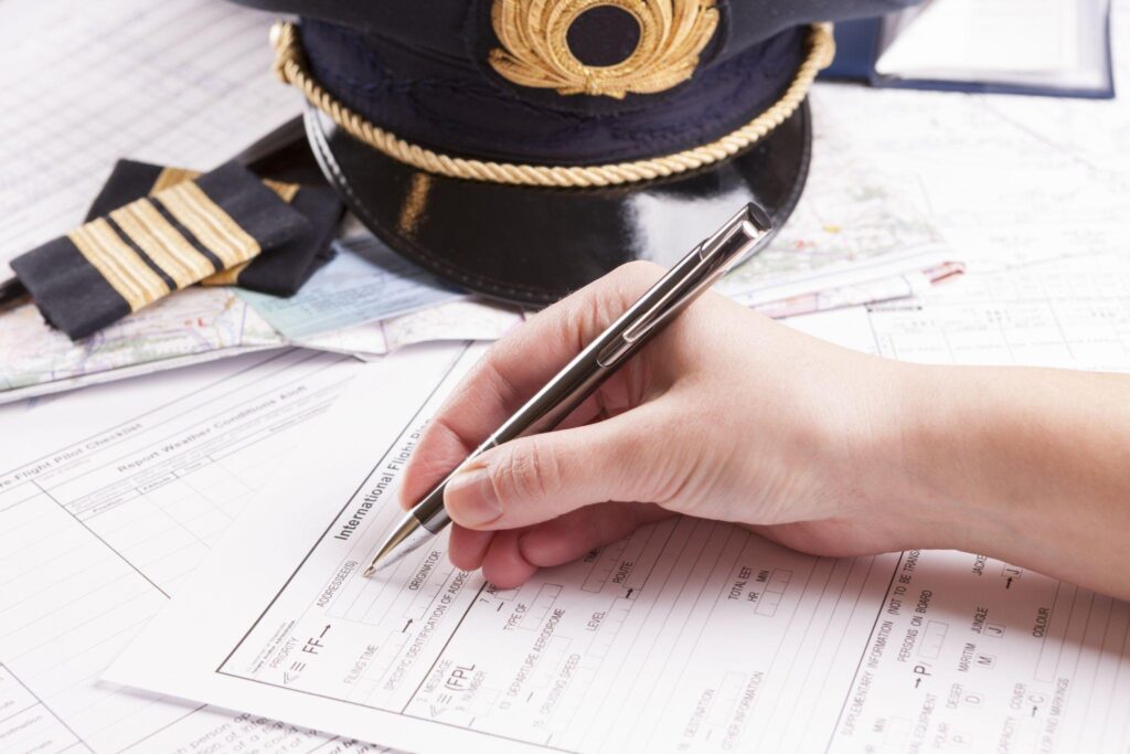 an airplane pilot hand filling in an flight plan with equipment including hat, epaulettes and other documents in background