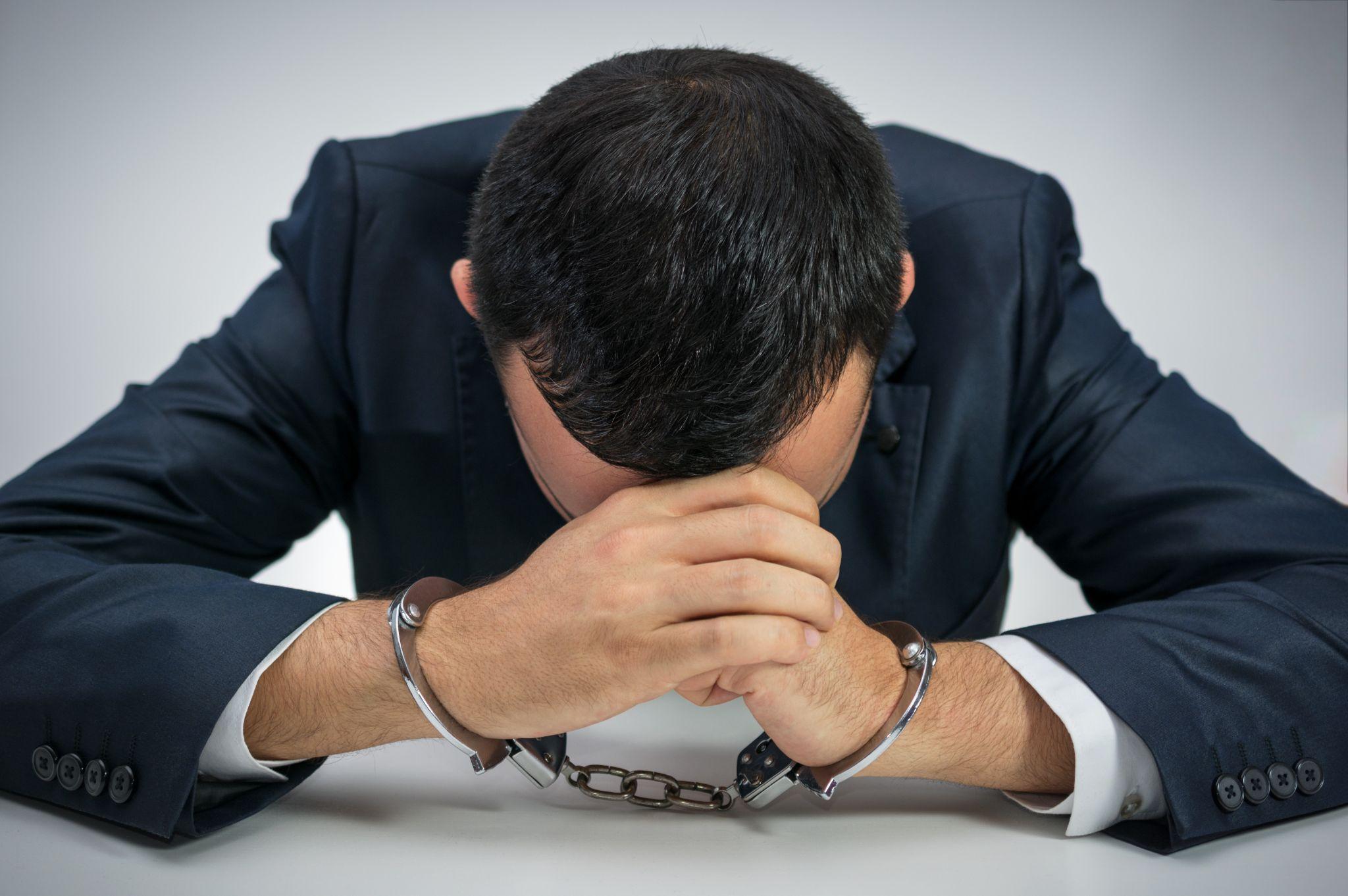 Young businessman handcuffed. He is sitting at a table with face between his palms showing guilt and frustration
