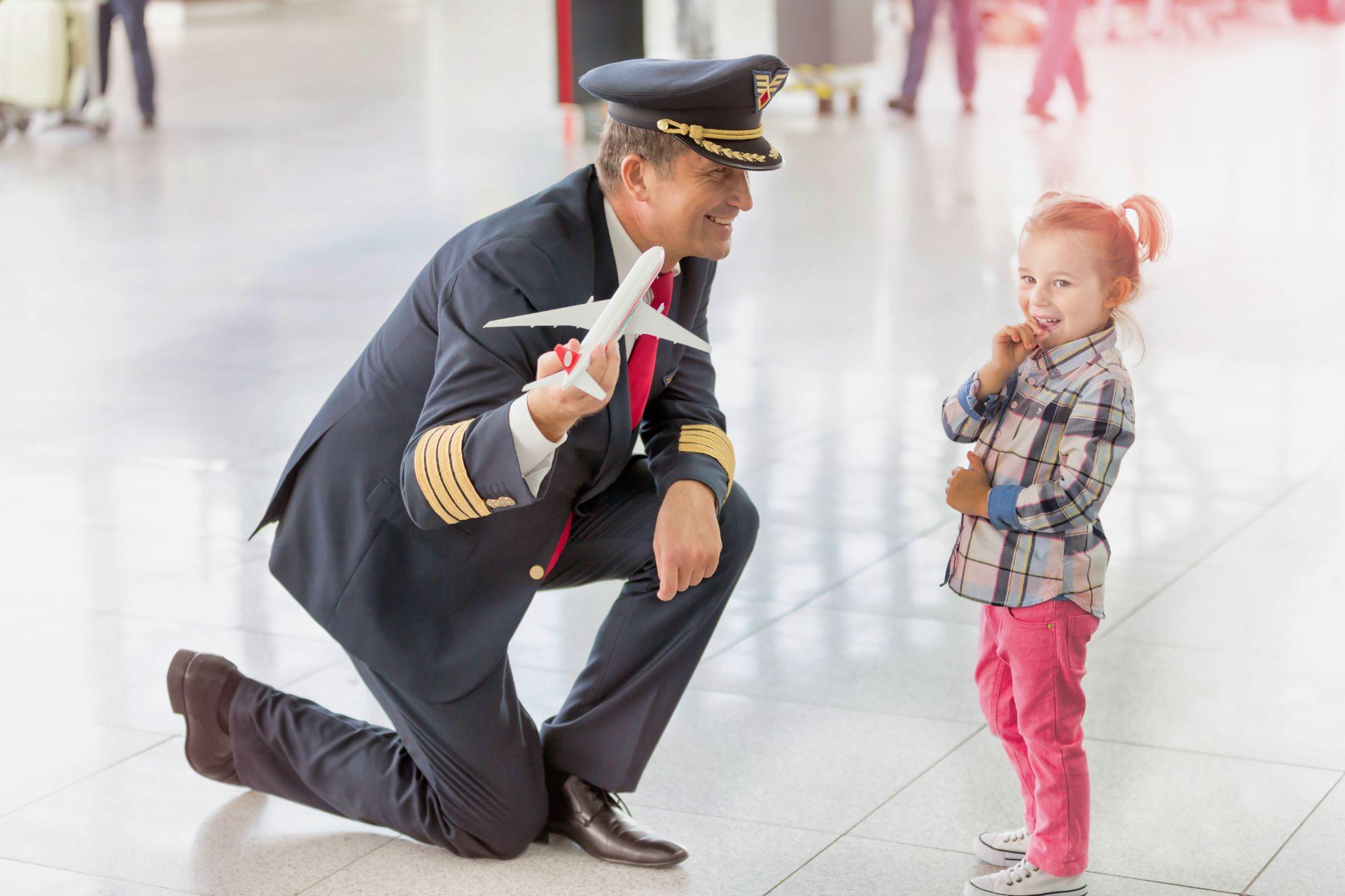 Portrait of mature pilot holding airplane toy while playing with cute little girl in airport.